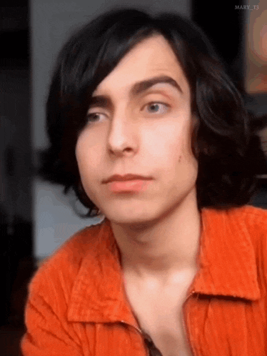 a young man with long black hair is wearing a red shirt and looking at the camera .