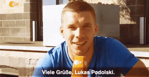 a man in a blue shirt is smiling and holding a microphone with the words viele grube lukas podolski below him