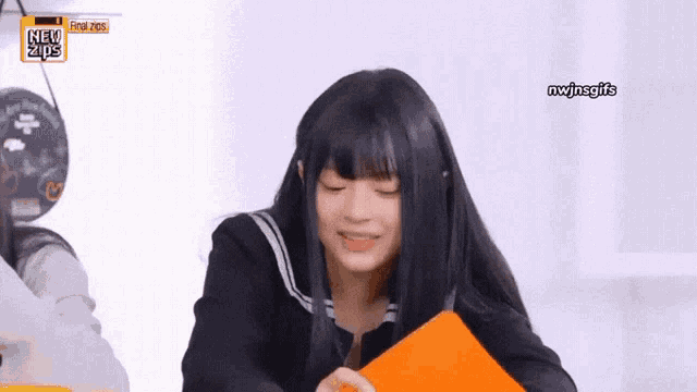 a girl with long black hair is smiling and holding an orange piece of paper in front of a sign that says new zips