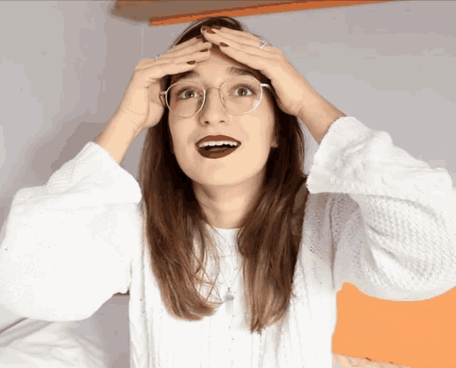 a woman wearing glasses and a white sweater has her hands on her forehead
