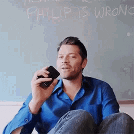 a man is sitting on the floor talking on a cell phone in front of a blackboard that says philip is wrong .