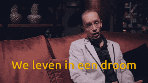 a man in a white suit sits on a red couch with the words we leven in een droom above him