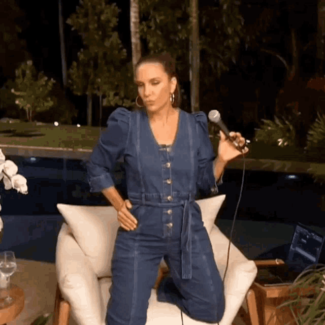 a woman in a denim jumpsuit is singing into a microphone while sitting in a chair