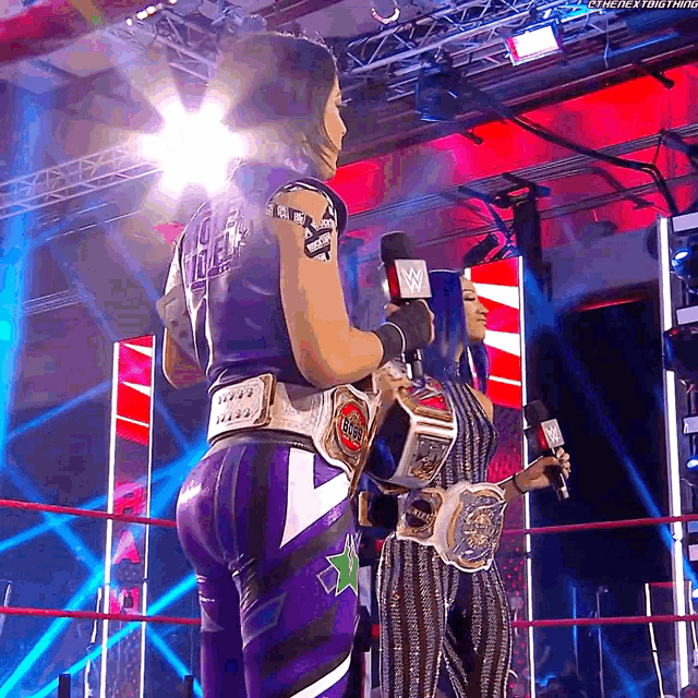 two women are standing in a wrestling ring holding championship belts and talking into microphones ..