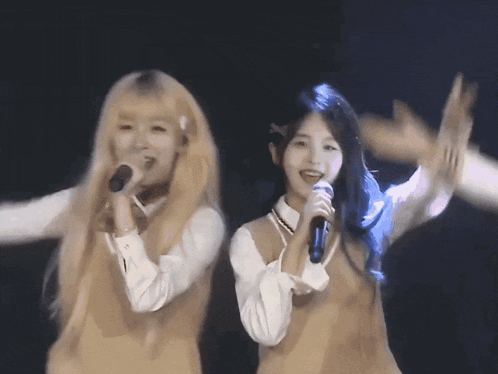 two girls singing into microphones on a stage with their arms outstretched