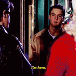 a man in a brown jacket is standing next to a woman in a red dress and says i 'm here .