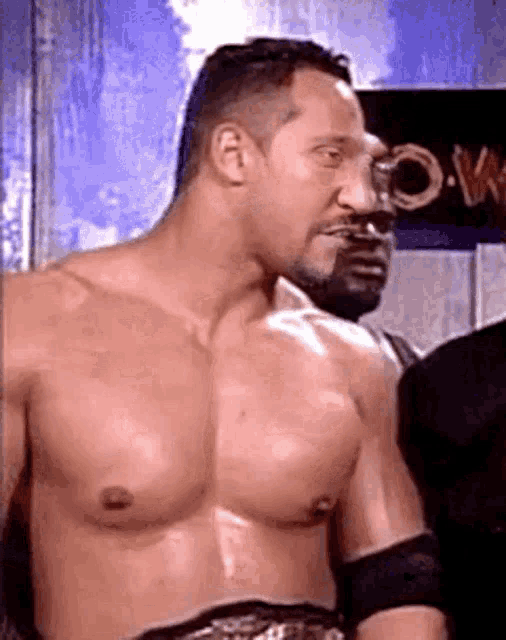 a shirtless wrestler is standing next to another wrestler in a locker room .