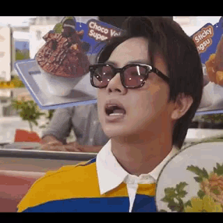 a man wearing sunglasses and a yellow and blue shirt is sitting in front of a display of food .