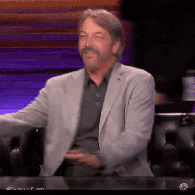 a man in a suit is sitting at a table with his hands in his pockets and laughing .