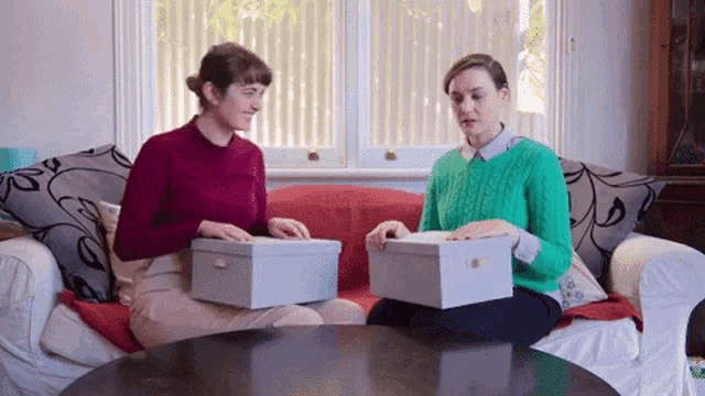 two women are sitting on a couch looking at boxes