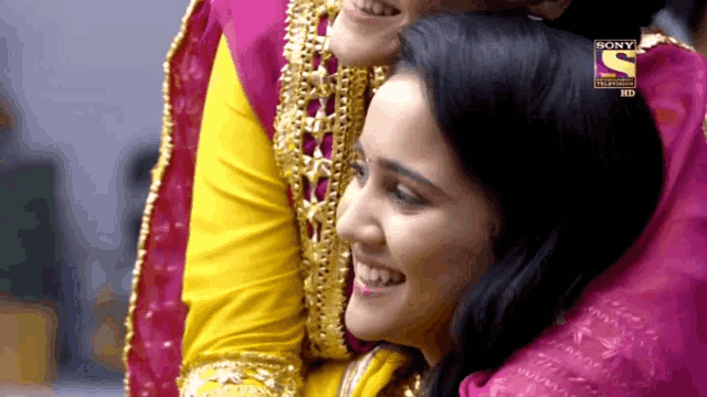 a woman in a yellow and pink dress is hugging another woman in a pink and yellow dress with a sony logo on her head