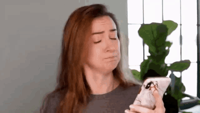 a woman is making a funny face while holding a cell phone in front of a plant .