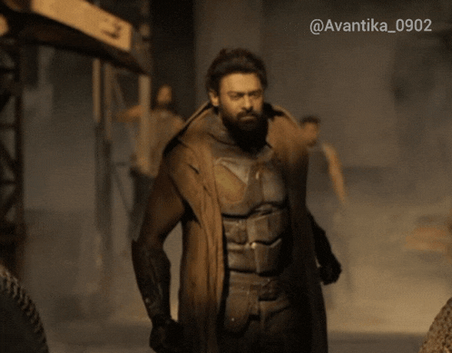 a man in a trench coat stands in front of a sign that says ' avantika_0902 ' on it