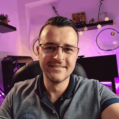 a man wearing glasses is smiling in front of a purple wall with a picture of a clown on it