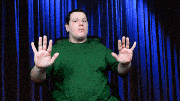 a man in a green shirt sitting in front of a blue curtain with his hands outstretched