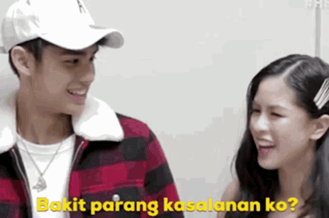 a man and a woman are standing next to each other and smiling . the man is wearing a baseball cap .
