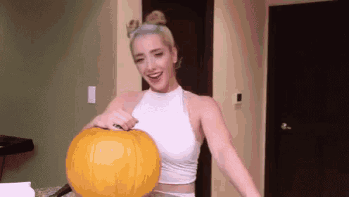 a woman in a white tank top is holding a large pumpkin .