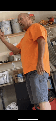 a man in an orange shirt and blue shorts is standing in front of a shelf in a garage .
