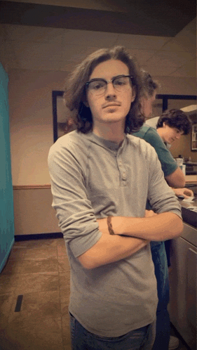 a young man wearing glasses and a gray shirt stands with his arms crossed