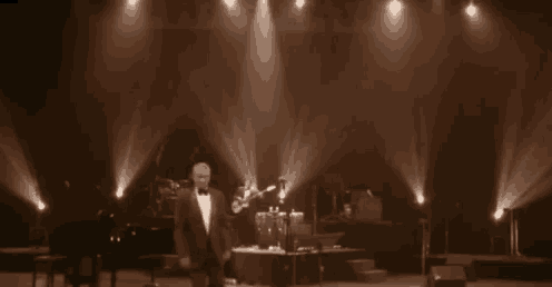 a man is standing on a stage in front of a piano while a band plays behind him .