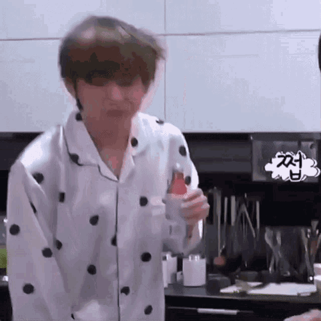 a man in a white shirt with black polka dots is standing in a kitchen holding a bottle .
