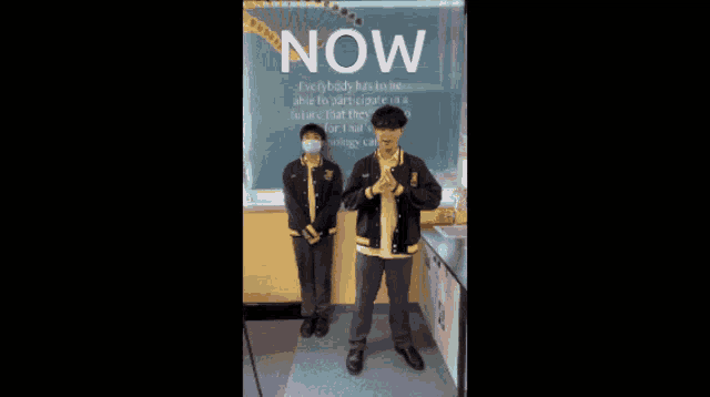 two boys are standing in front of a sign that says " now "
