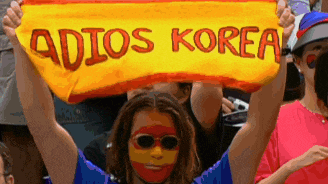 a woman with spanish paint on her face holds up a yellow banner that says adios korea
