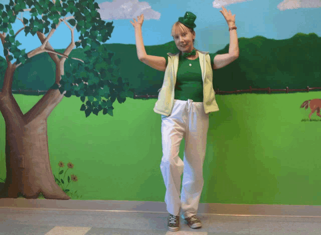 a woman in a green shirt and white pants is standing in front of a painting of a tree