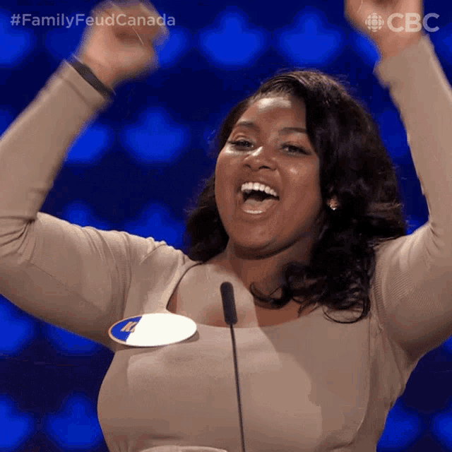 a woman stands in front of a microphone with her arms in the air and the words family feud canada on the bottom