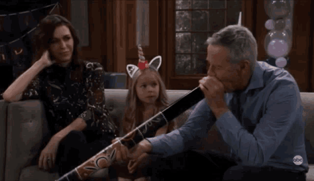 a little girl with unicorn ears sits on a couch next to a man holding a long stick