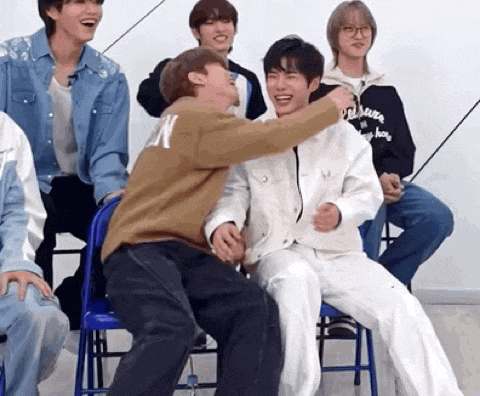 a group of young men are sitting in chairs and laughing . one of the men is hugging another man .