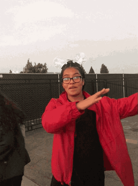 a girl wearing glasses and a red jacket stands in front of a black fence
