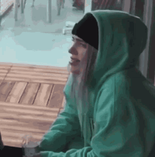 a woman wearing a green hoodie and a black beanie is sitting on a wooden floor .