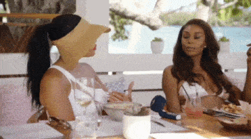 two women are sitting at a table with glasses of wine and food . one of the women is wearing a mask on her face .