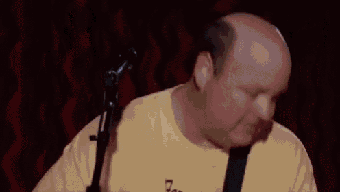 a man wearing a yellow shirt that says farmer 's daughter is singing into a microphone while holding a guitar .