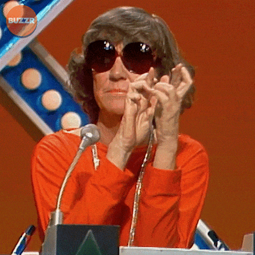 a woman wearing sunglasses is clapping her hands in front of a microphone with the buzzr logo behind her