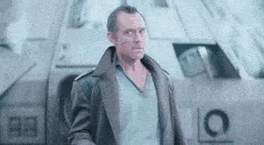 a man in a trench coat is standing in front of a large vehicle .