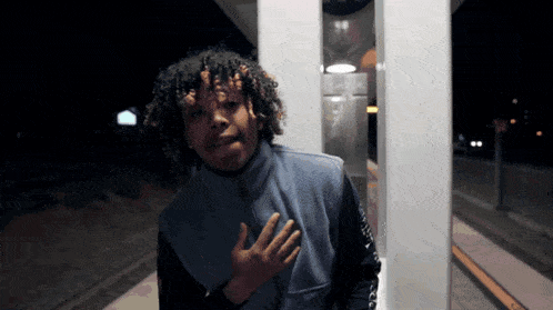 a man with curly hair is standing in front of a telephone booth with his hand on his chest