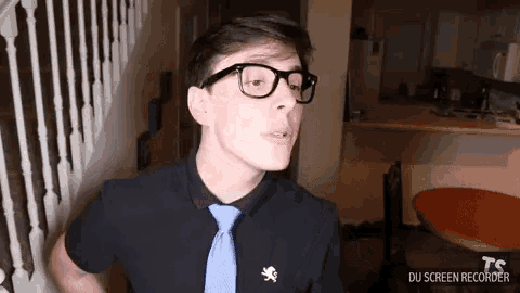 a young man wearing glasses , a black shirt and a blue tie is standing in a room .