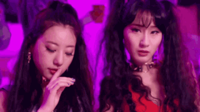 two women are standing next to each other in front of a purple background . one of the women is covering her mouth with her hand .