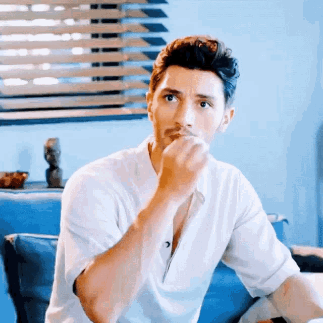 a man in a white shirt is sitting on a blue couch with his hand on his chin