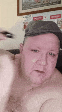 a shirtless man wearing a hat and a necklace is standing in front of a wall with a sign that says become