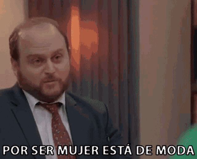 a man in a suit and tie is standing in front of a window and says `` por ser mujer esta de moda '' .