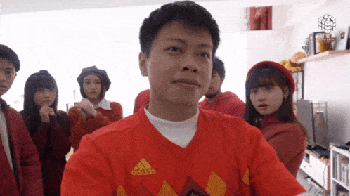a man wearing a red adidas shirt stands in a room with other people