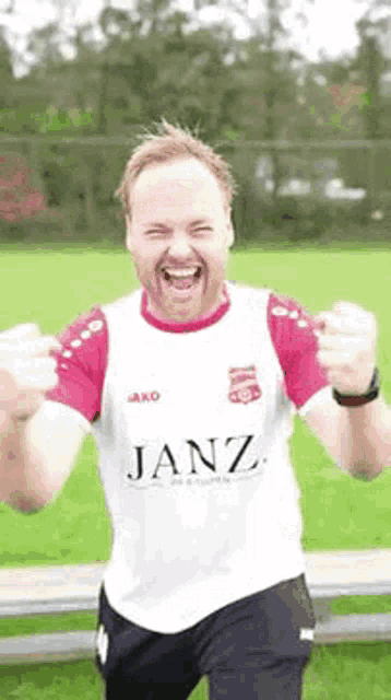a man wearing a janz shirt is standing in a field with his hands in the air .