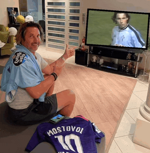 a man sitting in front of a television with a purple jersey that says mostovoi on it