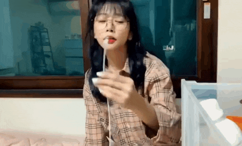 a woman wearing glasses and a plaid shirt is sitting on a bed holding a marshmallow in her mouth .