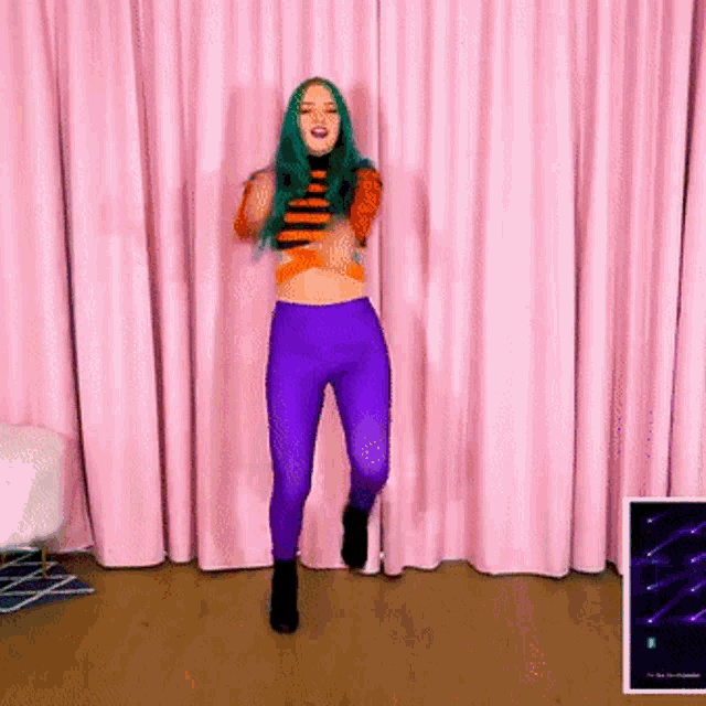 a girl with green hair and purple pants is dancing in front of a pink curtain