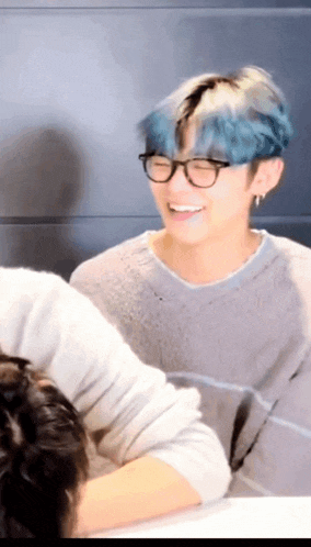 a man with blue hair is wearing glasses and a grey sweater