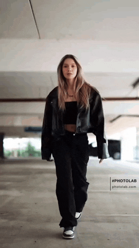 a woman in a black jacket and black pants is walking in a parking garage